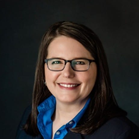 A woman with straight brown hair, wearing glasses, a blue collared shirt, and a dark blazer, smiles in front of a dark background.