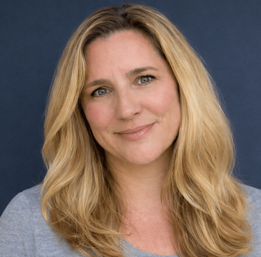A woman with long blonde hair and a grey shirt stands in front of a solid dark blue background, smiling softly at the camera.