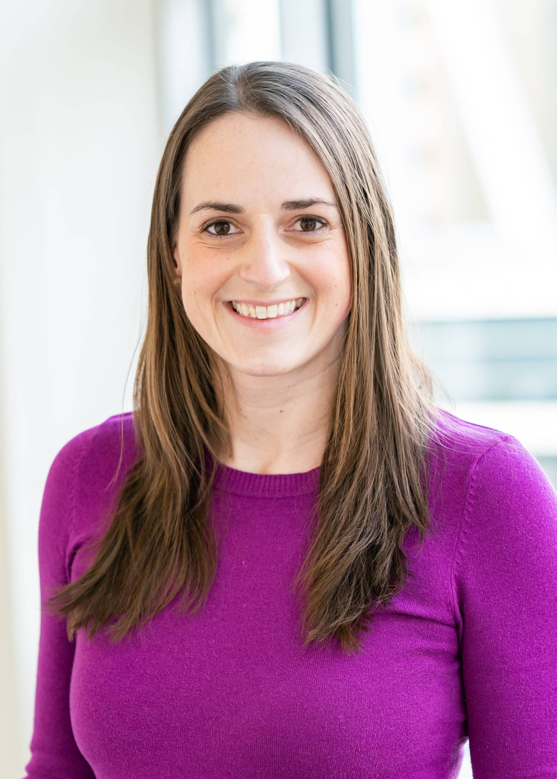 A woman with straight brown hair, wearing a purple sweater, smiles while standing indoors in front of bright windows—reflecting the positive impact of AI patient readiness and cancellation reduction in healthcare settings.