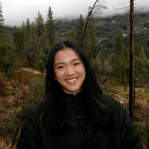 A young woman wearing a black jacket smiles at the camera while standing in a forested area with mountains and low clouds in the background.