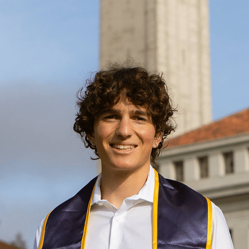 A person wearing a graduation sash and white shirt stands outdoors in front of a tall clock tower and a building with a red roof.