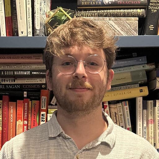 A man with glasses and a beard stands in front of a bookshelf filled with various books.