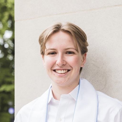 A person with short, light brown hair smiles at the camera, wearing a white collared shirt and standing outdoors near a light-colored stone wall.