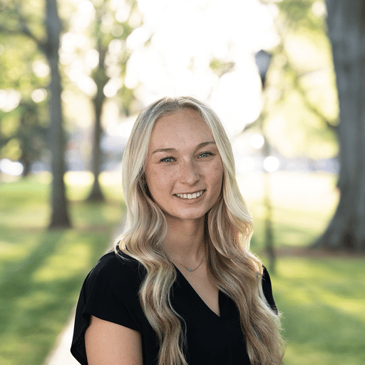 A young woman with long blonde hair and a black top stands outdoors in a park, smiling at the camera. Trees and sunlight fill the blurred background.