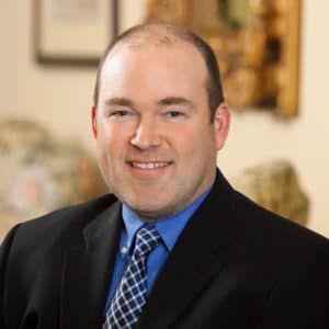A man in a suit and blue shirt with a checkered tie sits indoors, smiling at the camera. The background is softly blurred, showing framed art and patterned furniture.