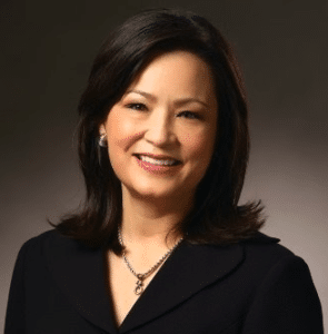 A woman with shoulder-length dark hair, wearing a black blazer, a necklace, and earrings, smiles at the camera against a plain, dark background.