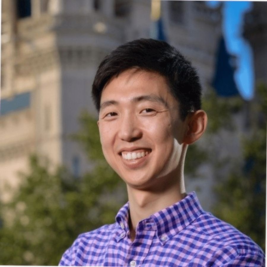 A man with short black hair, wearing a purple checkered shirt, smiles while standing outdoors with a blurred historic building in the background.