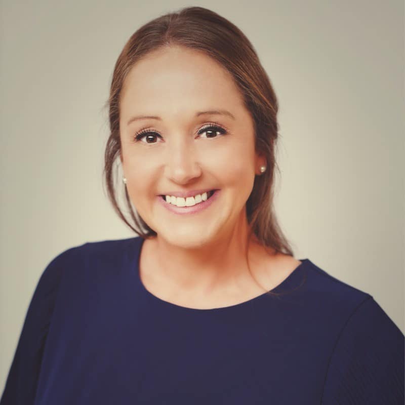 A woman with long brown hair and a navy blue top smiles at the camera against a plain, light-colored background.