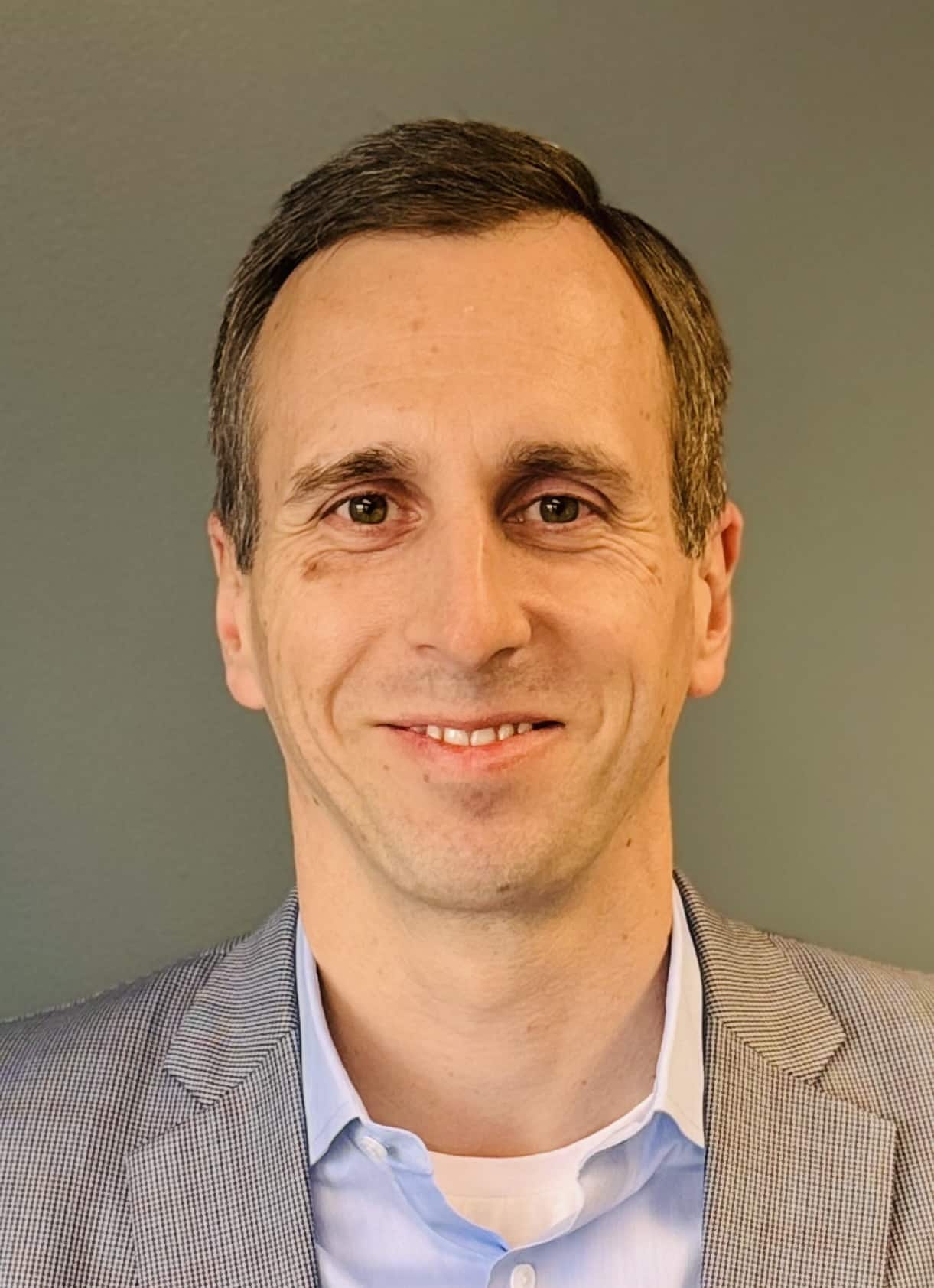 A man with short brown hair, wearing a light blue collared shirt and a gray blazer, smiles at the camera against a plain background, ready to unlock capacity with intelligent workflows.