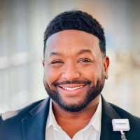 A man in a suit jacket and white shirt smiles at the camera, proudly wearing a name badge. The blurred background hints at a conference focused on building resilient sustainable health systems.