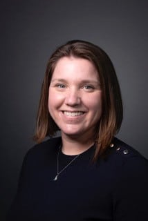 A woman with straight brown hair, wearing a dark top and necklace, poses and smiles in front of a plain dark background.