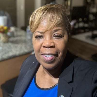 A woman with short, light brown hair smiles at the camera, wearing a blue top and a black blazer in a kitchen setting—her confident look reflecting expertise in infusion center workforce management.