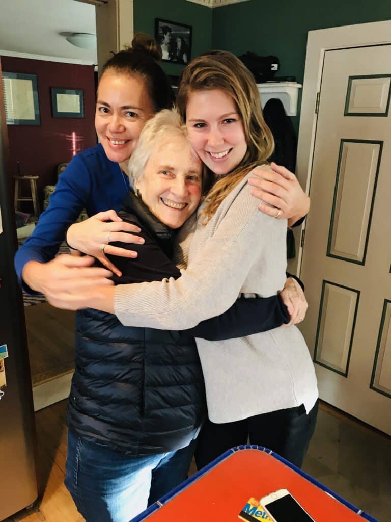 Three women stand indoors, smiling and hugging each other in front of a door. A blue table with a smartphone and mail is visible in the foreground.