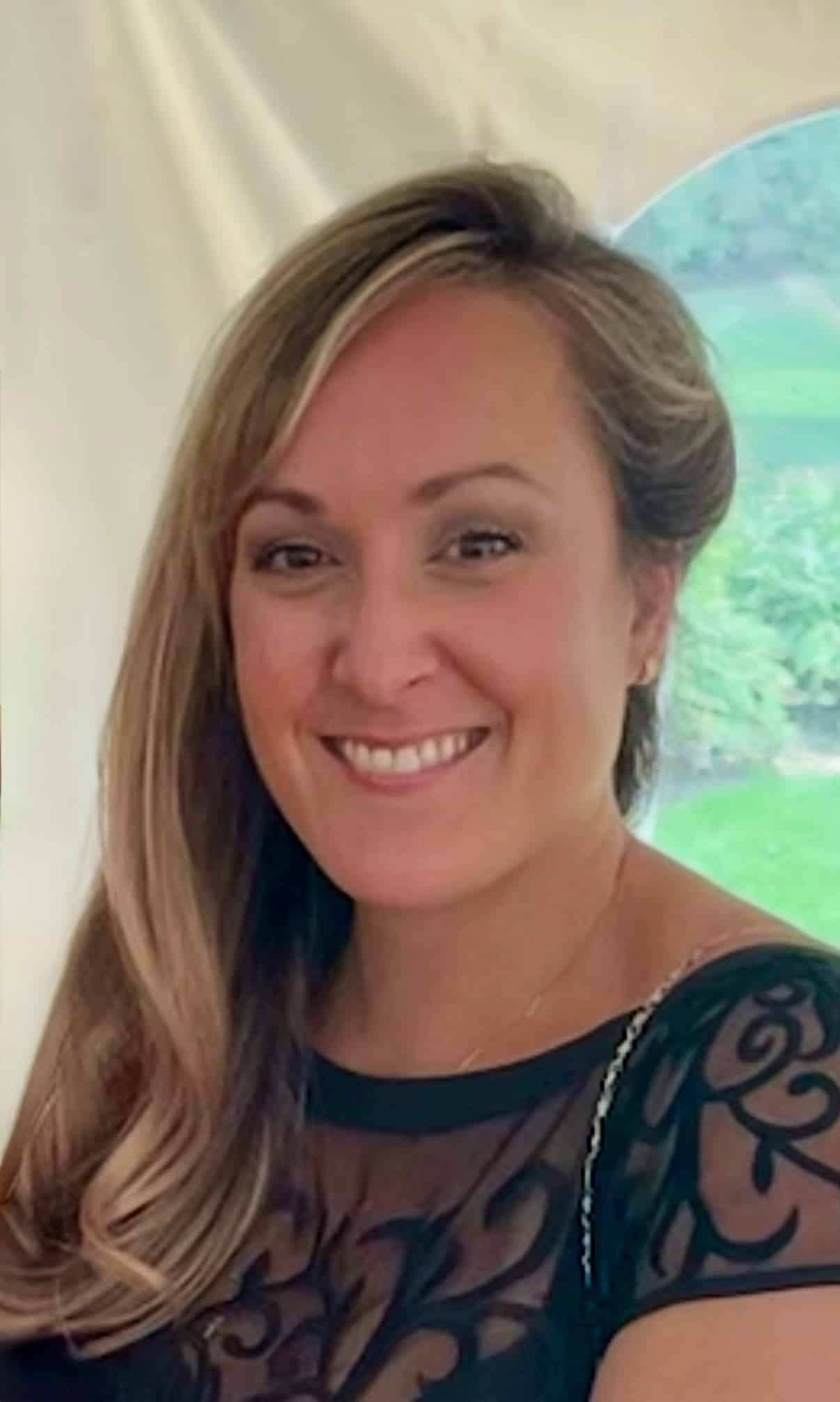 A woman with long, light brown hair and a black patterned dress smiles at the camera in an outdoor setting.