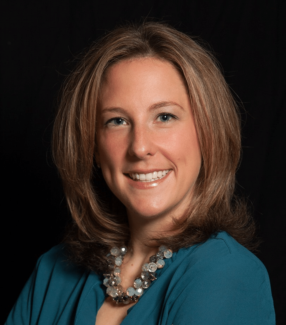 A woman with shoulder-length light brown hair, wearing a teal top and a beaded necklace, smiles in front of a black background.