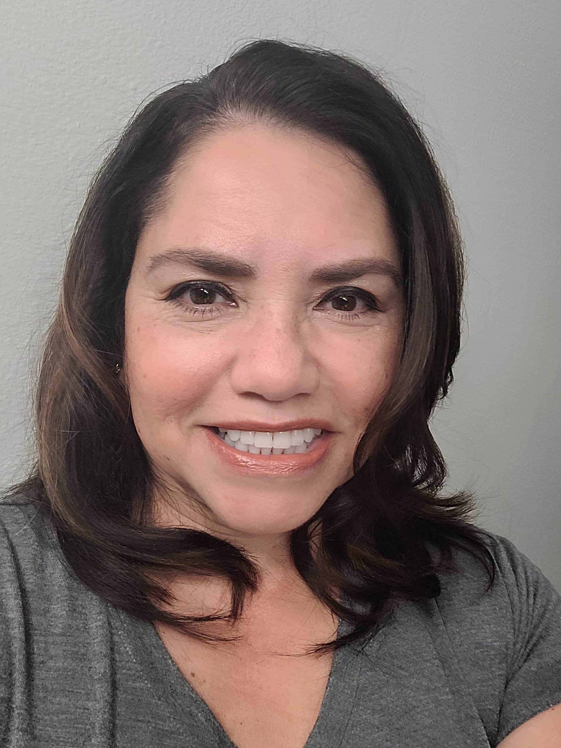 A woman with shoulder-length dark hair and a gray shirt smiles at the camera against a plain light-colored background, reflecting the capacity to unlock potential with intelligent workflows.