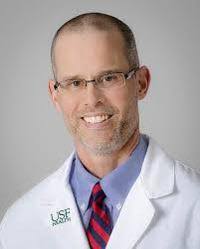 A man wearing glasses, a white lab coat with a USF Health logo, a blue shirt, and a red-striped tie, smiling at the camera against a plain background—reflecting dedication to resilient sustainable health systems.