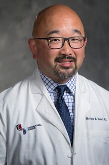 A man wearing glasses, a white medical coat, a checkered shirt, and a blue tie stands in front of a neutral background, ready to unlock capacity with intelligent workflows.