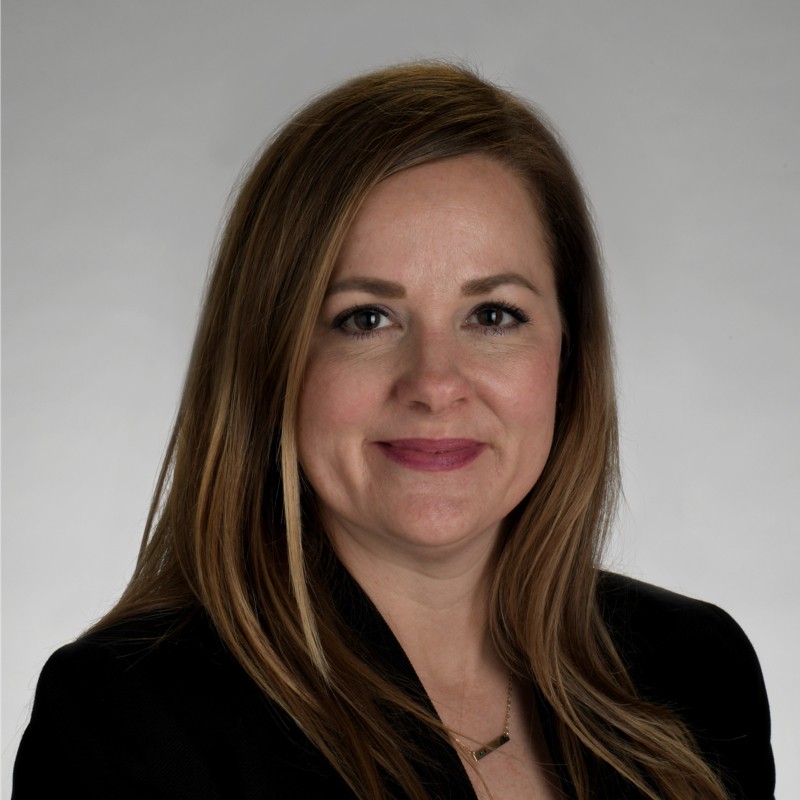 A woman with long light brown hair, wearing a black blazer and a necklace, poses for a professional headshot in front of a plain light gray background, ready to unlock her full capacity with intelligent workflows.