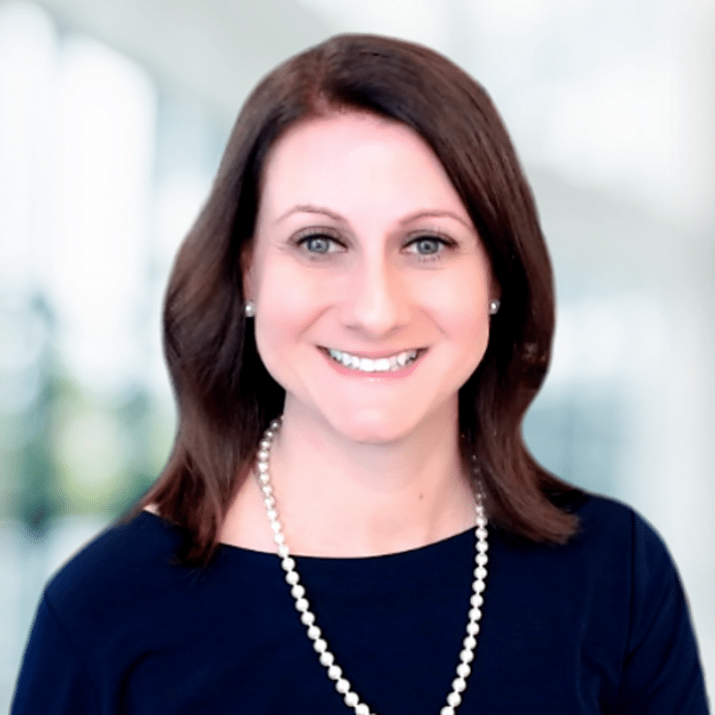 A woman with straight brown hair, wearing a dark top and a pearl necklace, smiles in a brightly lit, blurred indoor setting.