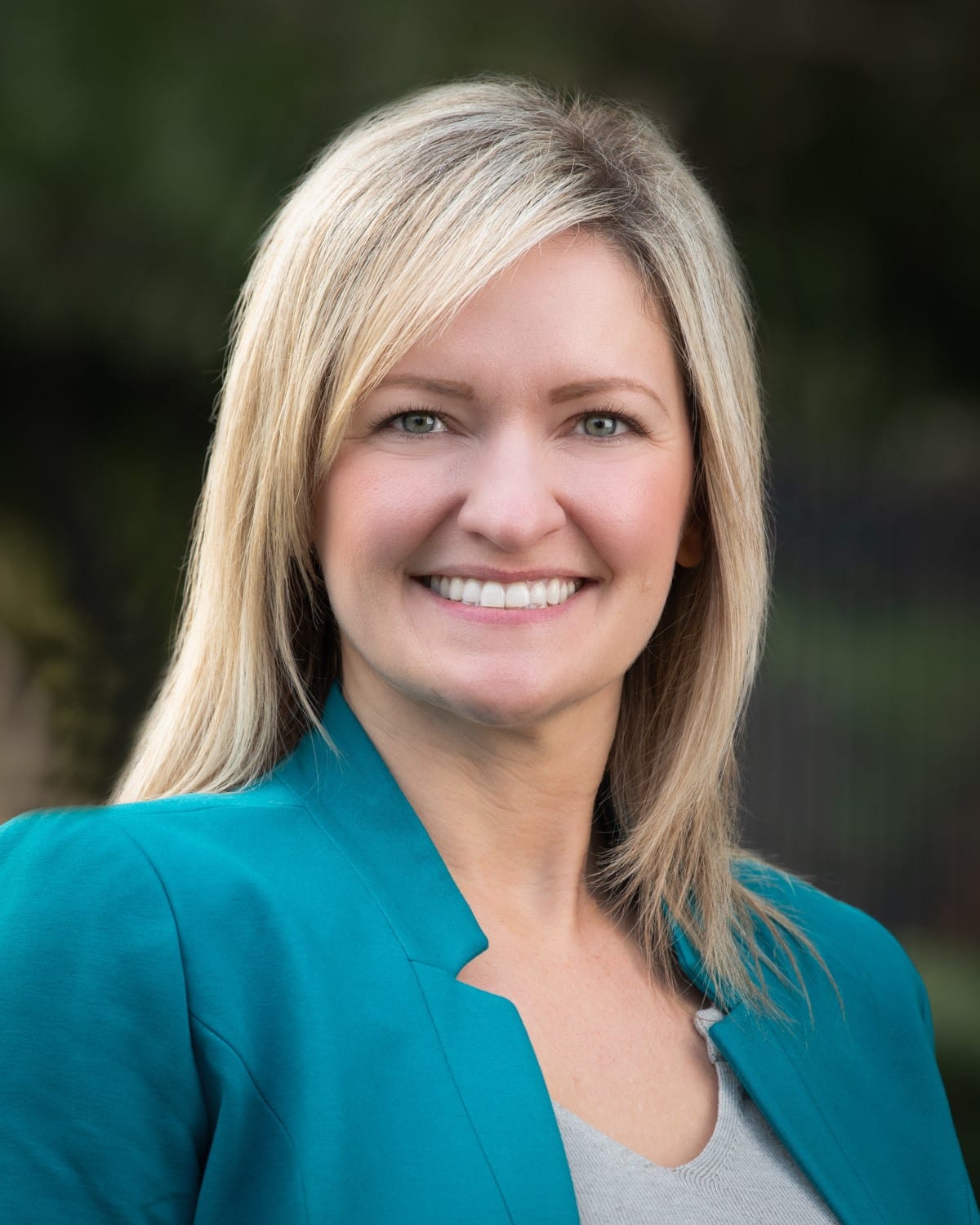 A woman with straight blonde hair wearing a teal blazer and light top smiles at the camera, representing the professionalism found in infusion center workforce management, with a blurred outdoor background.