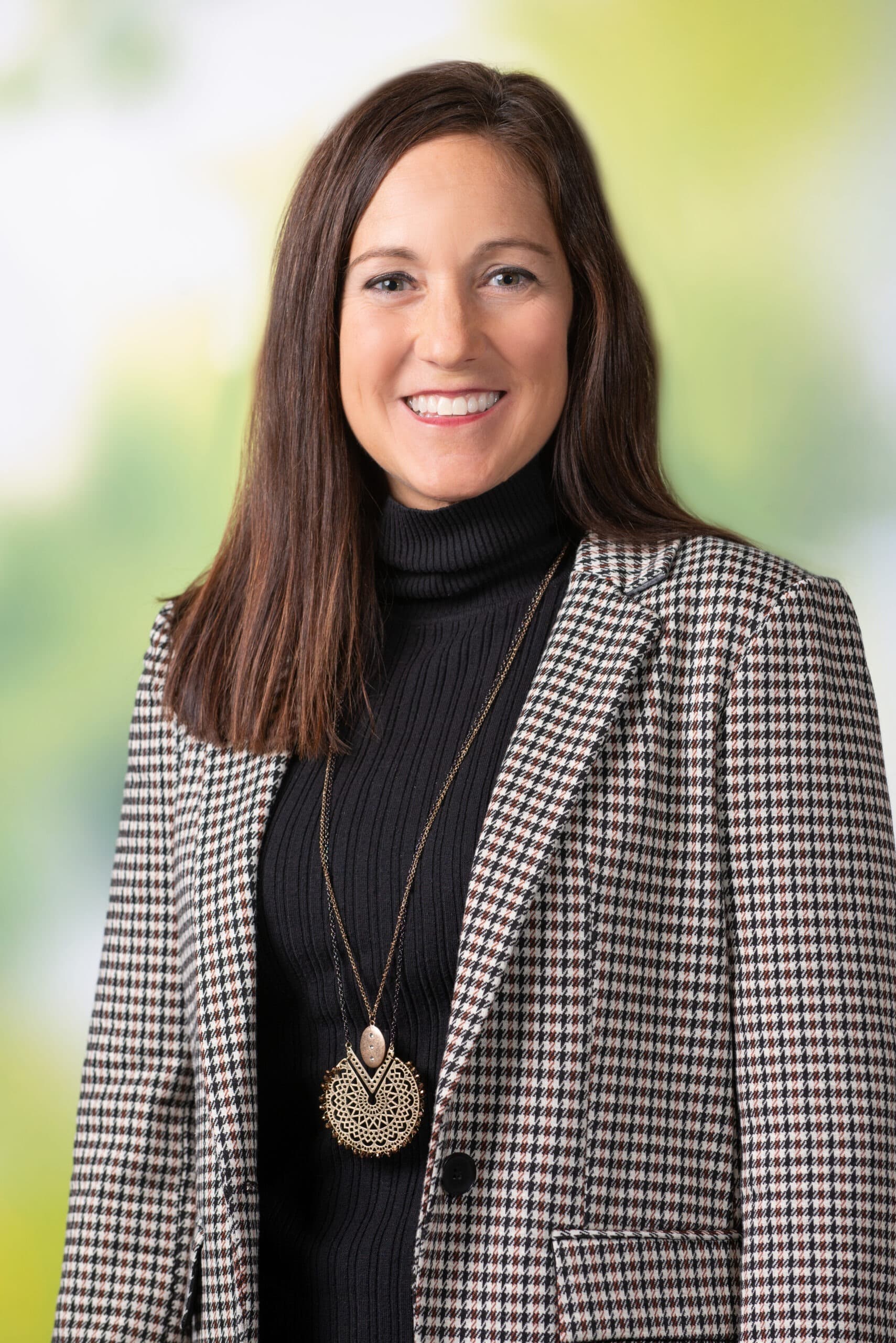 Woman with straight brown hair wearing a black turtleneck sweater, checked blazer, and a long pendant necklace, standing in front of a blurred green background—embodying the confidence seen in leaders of resilient sustainable health systems.