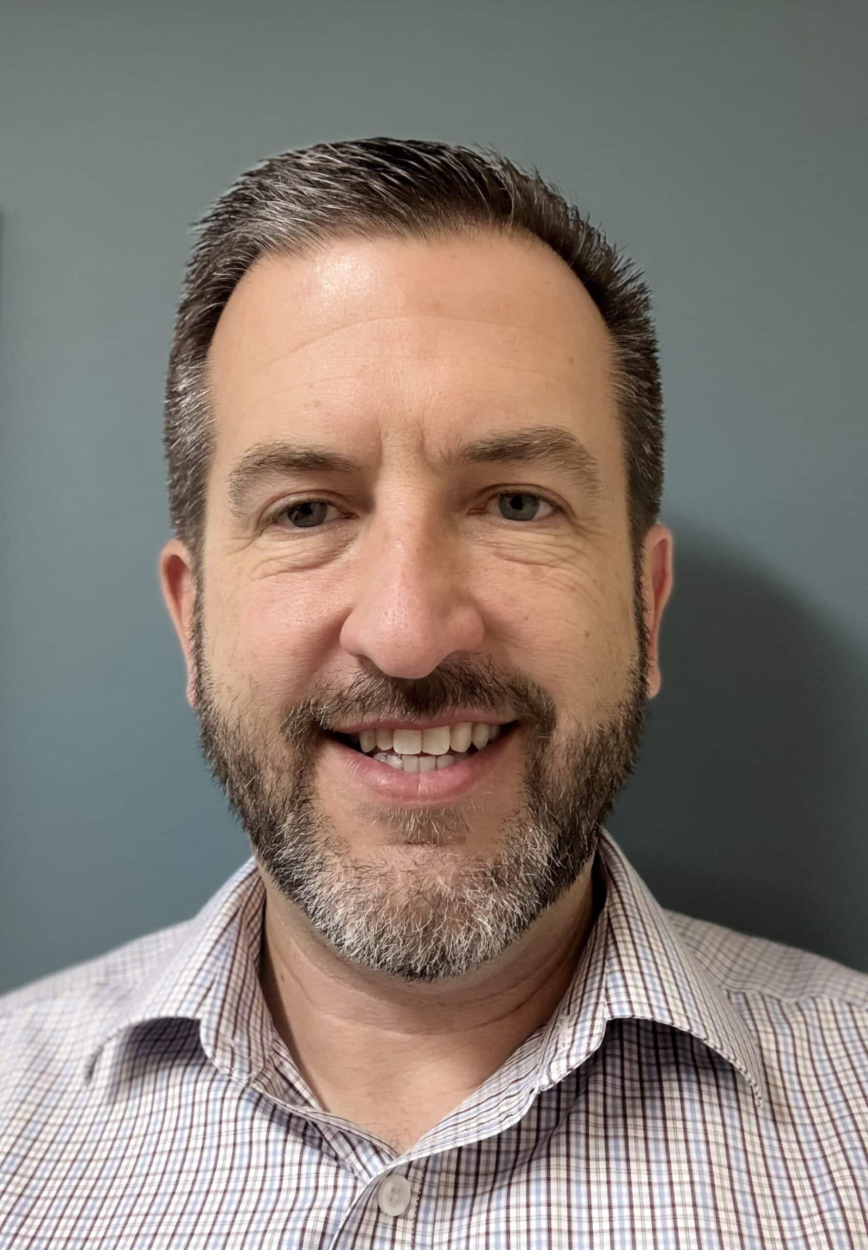 A man with short brown hair and a beard, wearing a light checkered shirt, smiles at the camera in front of a plain blue-gray background, embodying confidence in infusion center workforce management.
