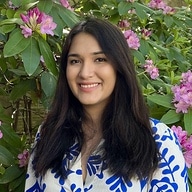 A woman with long dark hair, wearing a white and blue patterned top, stands in front of green bushes with pink flowers, smiling at the camera.