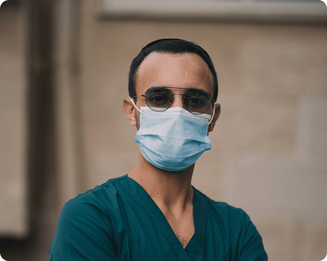 A doctor wearing medical scrubs, a face mask, and glasses stands with arms crossed, looking at the camera.