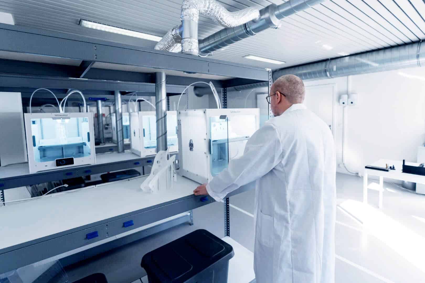 A person in a white lab coat operates a 3D printer in a clean, modern laboratory with shelves and ventilation ducts visible.