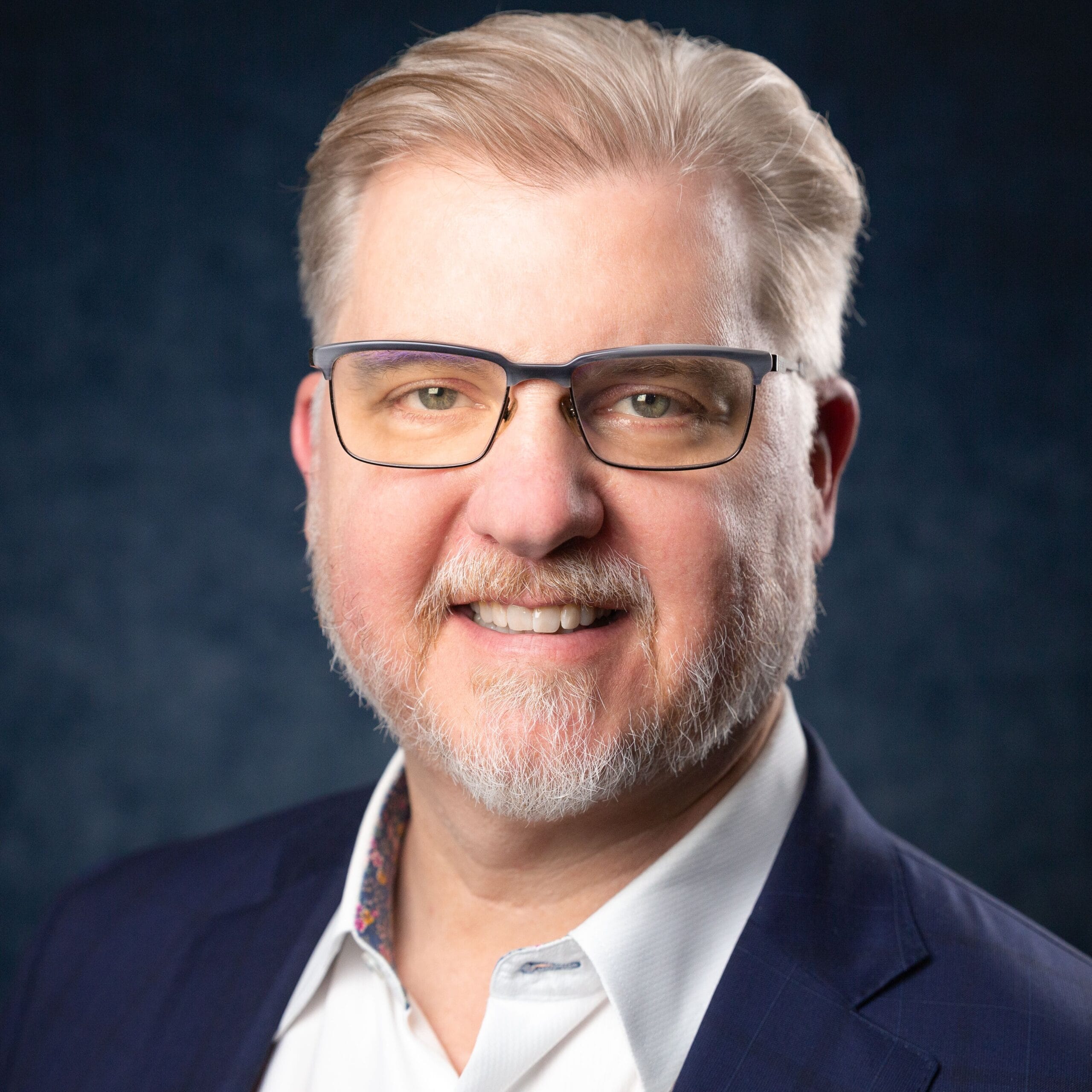A middle-aged man with light hair, beard, and glasses, wearing a dark suit jacket and white shirt, smiles in front of a dark blue background—an advocate for healthcare workforce optimization technology.