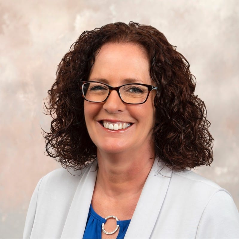 Woman with curly brown hair and glasses, wearing a white blazer over a blue top, smiling in front of a neutral background—perfect for representing innovation in surgical workflow.