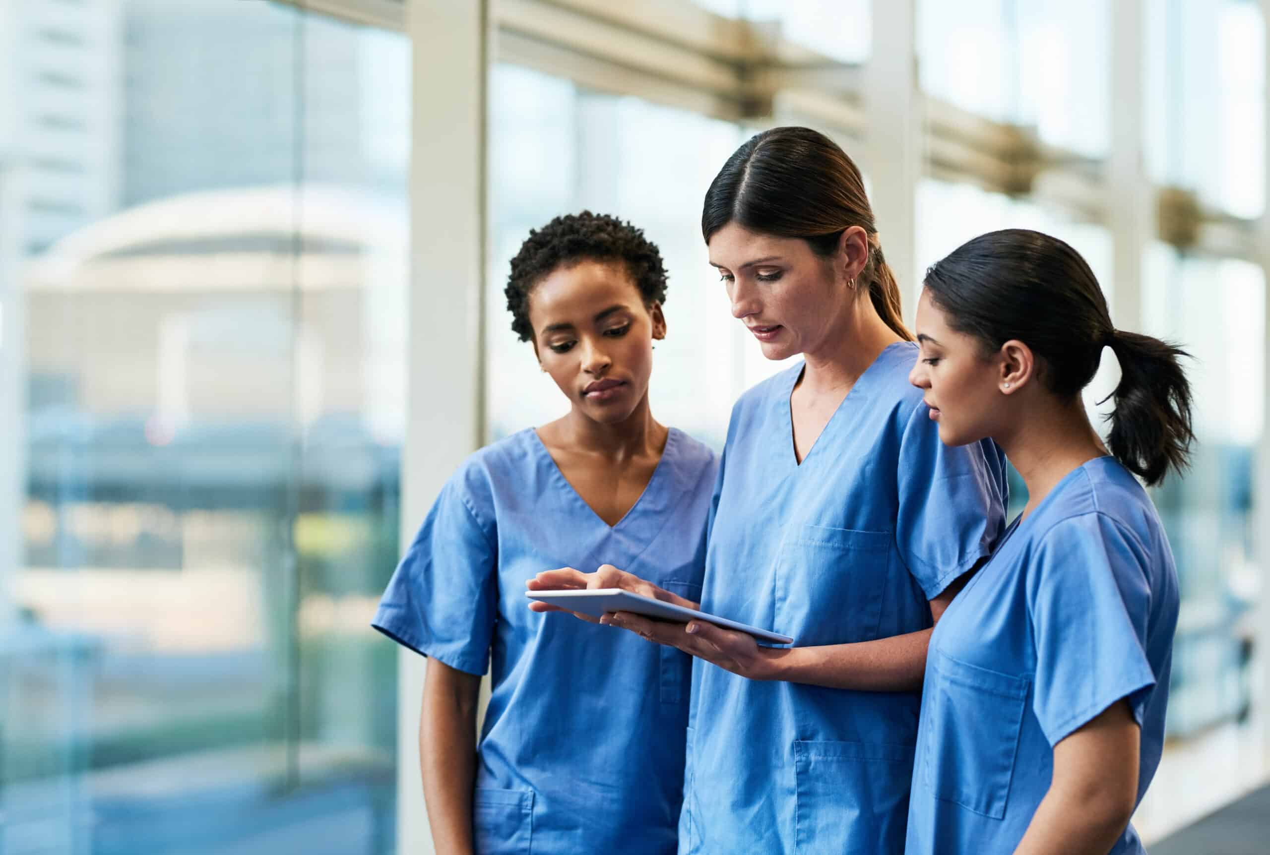 Three medical professionals in blue scrubs stand together indoors, looking at a clipboard and discussing.