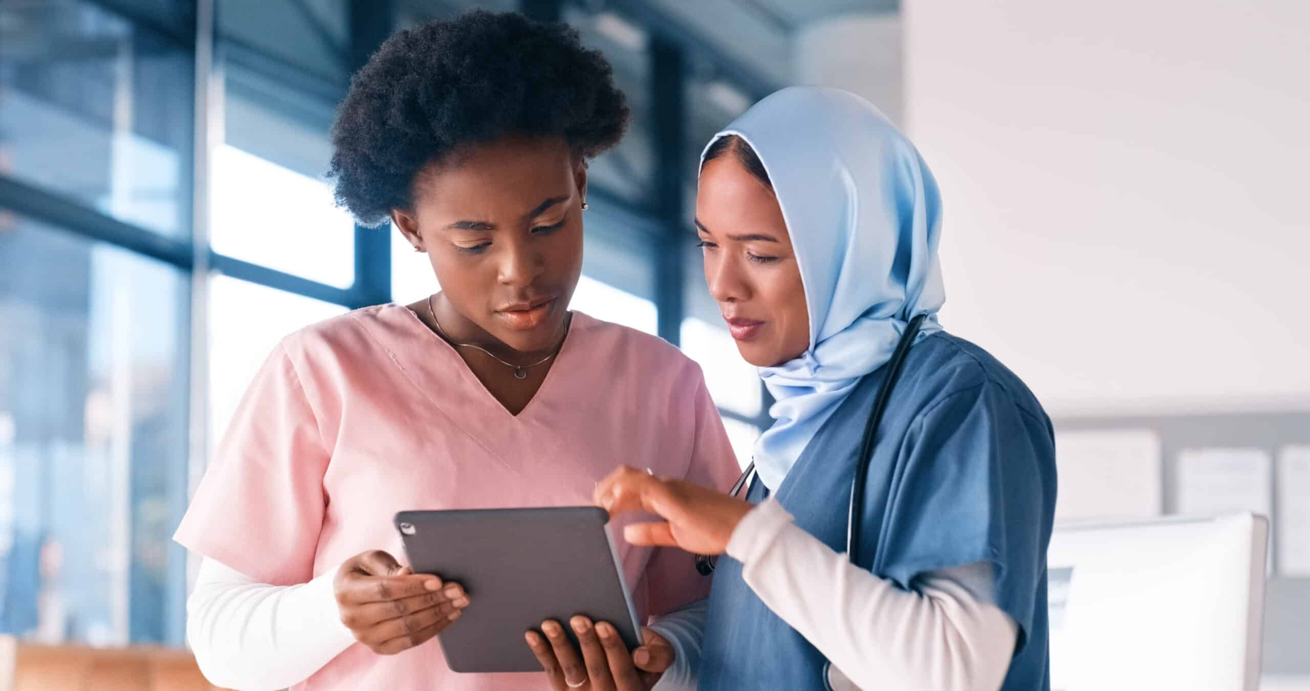 Two healthcare professionals review information on a digital tablet in a medical office setting, one wearing pink scrubs and the other in blue scrubs with a stethoscope and hijab.