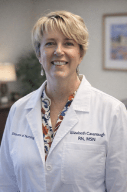 A woman in a white lab coat with “Director of Nursing” and “Elizabeth Cavanaugh, RN, MSN” embroidered, smiling in an office focused on infusion nurse scheduling optimization.