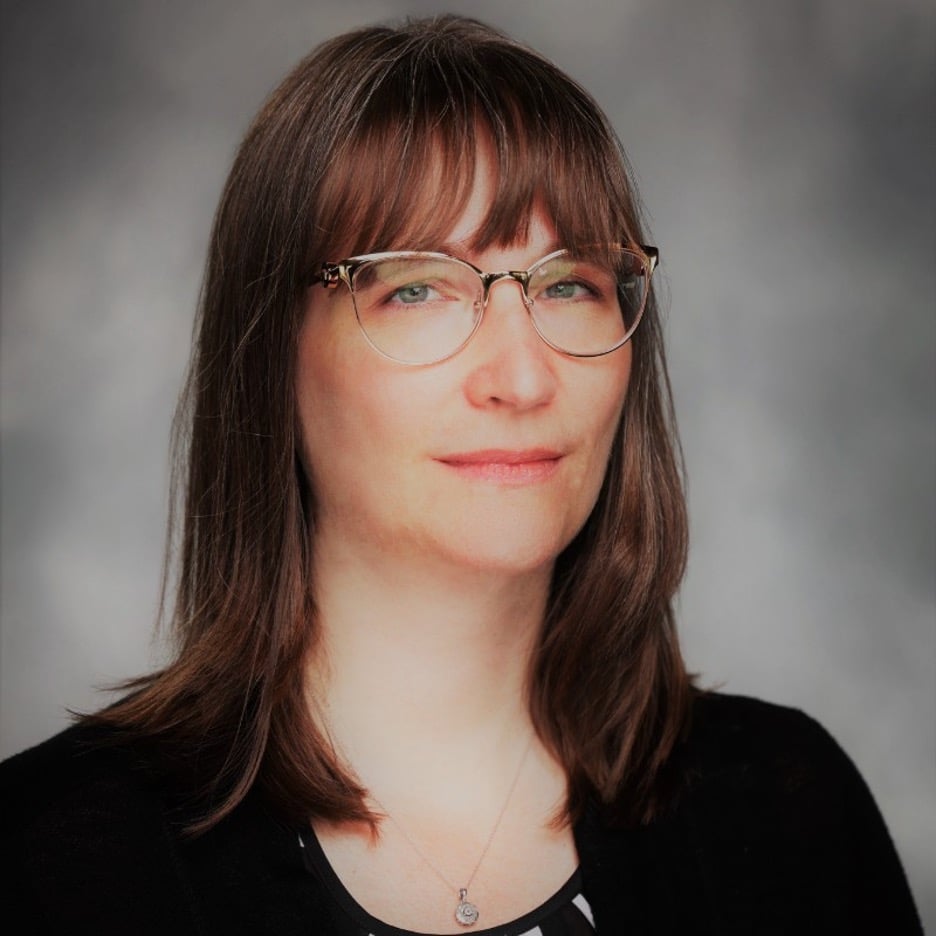 A woman with straight brown hair, glasses, and bangs, wearing a black top and necklace, poses in front of a gray background—ideal for infusion nurse scheduling optimization materials.