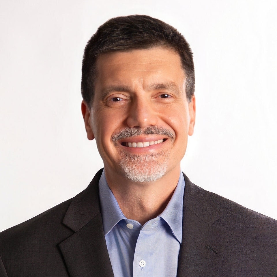Smiling middle-aged man with short dark hair and a goatee, wearing a dark suit jacket and light blue collared shirt, posed against a plain white background.