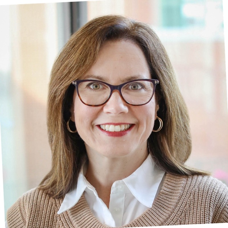 Woman with shoulder-length brown hair and glasses, wearing a light brown sweater over a white collared shirt, smiling in front of a blurred indoor OR utilization strategy background.