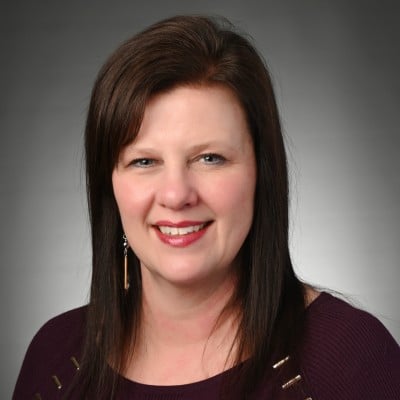 Woman with straight, shoulder-length dark brown hair wearing a maroon top, smiling at the camera against a plain gray background.