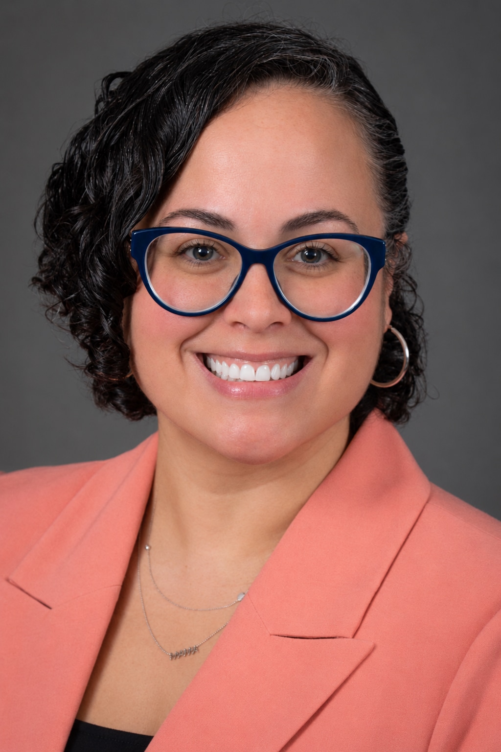 Smiling person with short curly hair, blue glasses, hoop earrings, and peach blazer—ready for scaling infusion operations—stands before a plain gray background.
