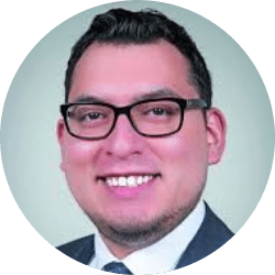 A man with short dark hair, glasses, and a trimmed beard, wearing a suit and tie, smiling at the camera against a light background.
